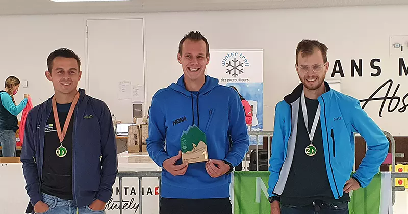 Drie glimlachende mannen staan op het podium. De man in het midden houdt een trofee vast en draagt een gouden medaille. De mannen links en rechts dragen een bronzen en een zilveren medaille. Alle drie dragen ze een sportjasje.